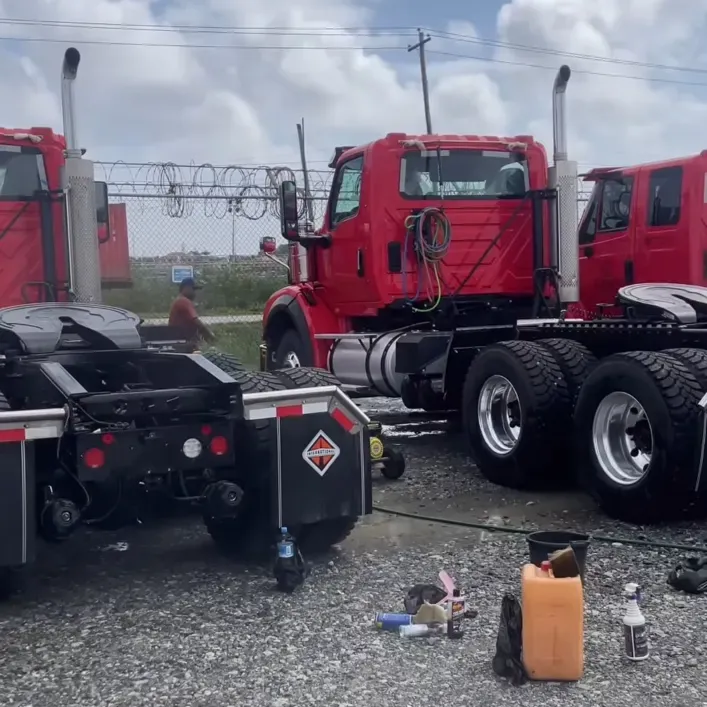 Red prime movers parked on a gravel fleet yard in Guyana during a CDCS on-site wash, with hoses and cleaning supplies on the ground