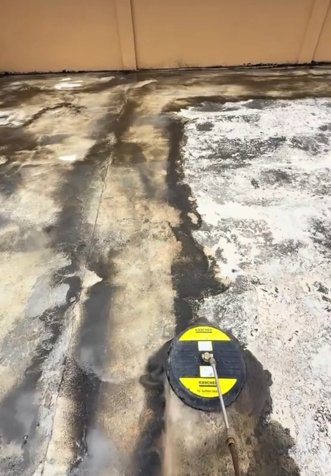 A rotary surface cleaner pressure washing a large concrete slab, with a cleaned strip showing against the grimy surface