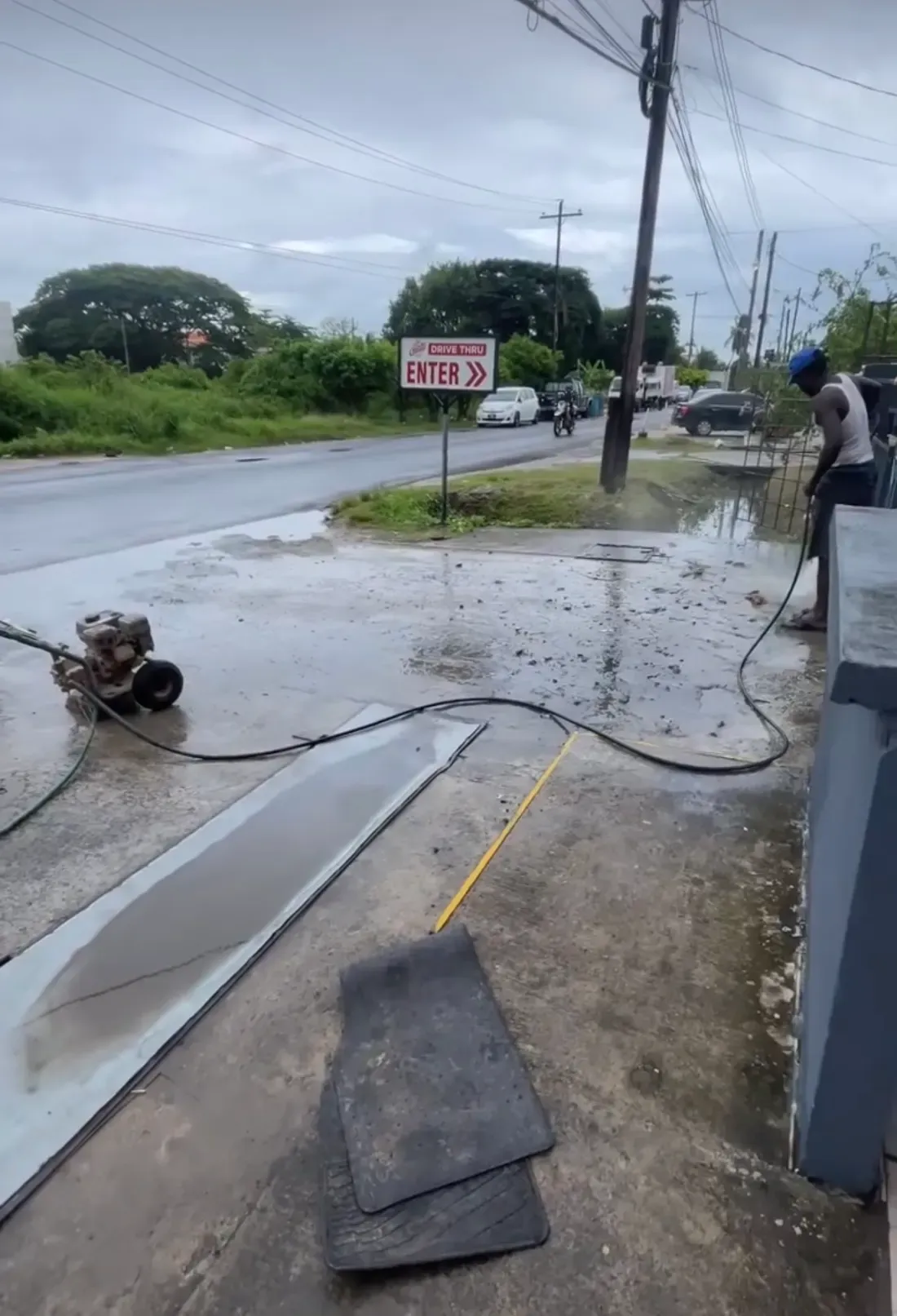 A CDCS worker pressure washing a boundary wall beside a road in Guyana, with a pressure washer on the ground